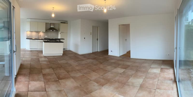 Empty room with tiled floor, white walls, and a kitchen area with cabinets and a sink visible in the background.
