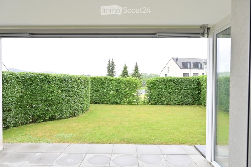 A well-maintained garden with a neatly trimmed hedge, a smooth lawn, and a paved patio area. The view includes a neighboring house and trees in the background.