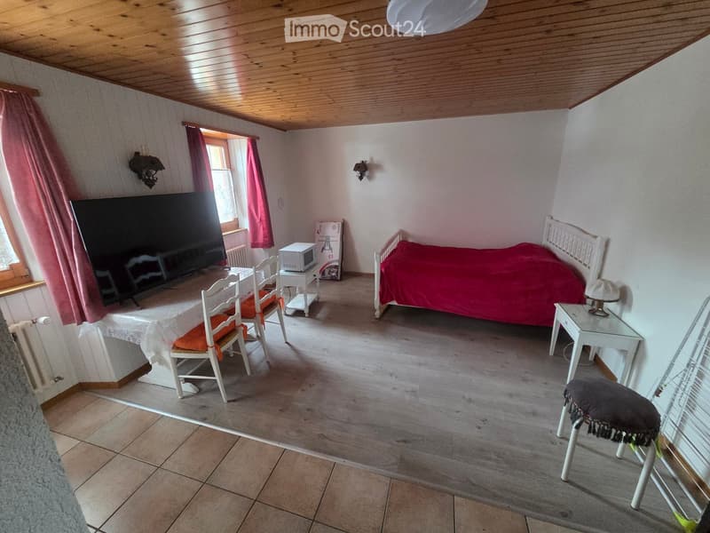 Furnished Room. Cozy bedroom with a bed, table, chairs, microwave, and a large screen. The room features wooden flooring and a slanted wooden ceiling.