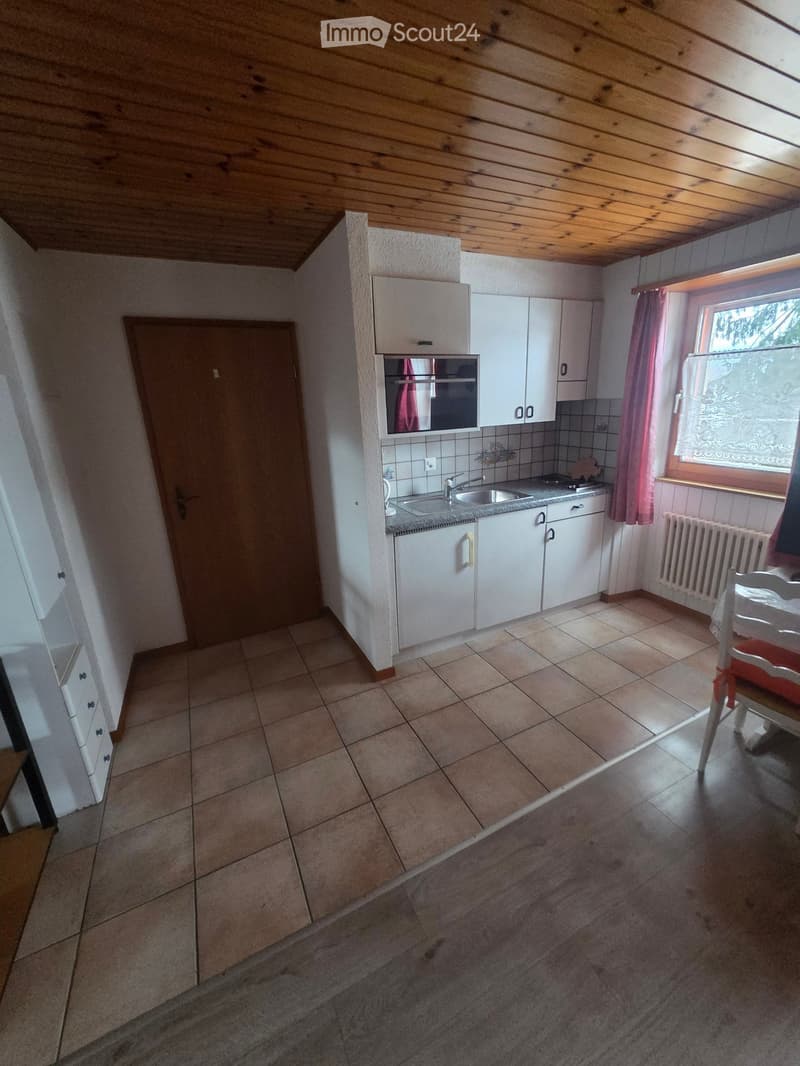 Furnished Room. The image shows a small, well-lit kitchen area with white cabinets, a sink, a stove, and a tiled floor. A wooden door is visible on the left, and a window with pink curtains is on the right. A small dining area with a chair and table is pa