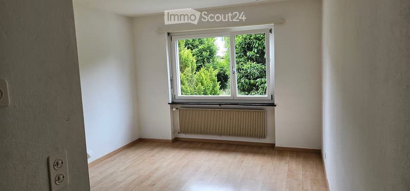 Empty room with wooden flooring, white walls, a window with a view of greenery, and a radiator below the window.