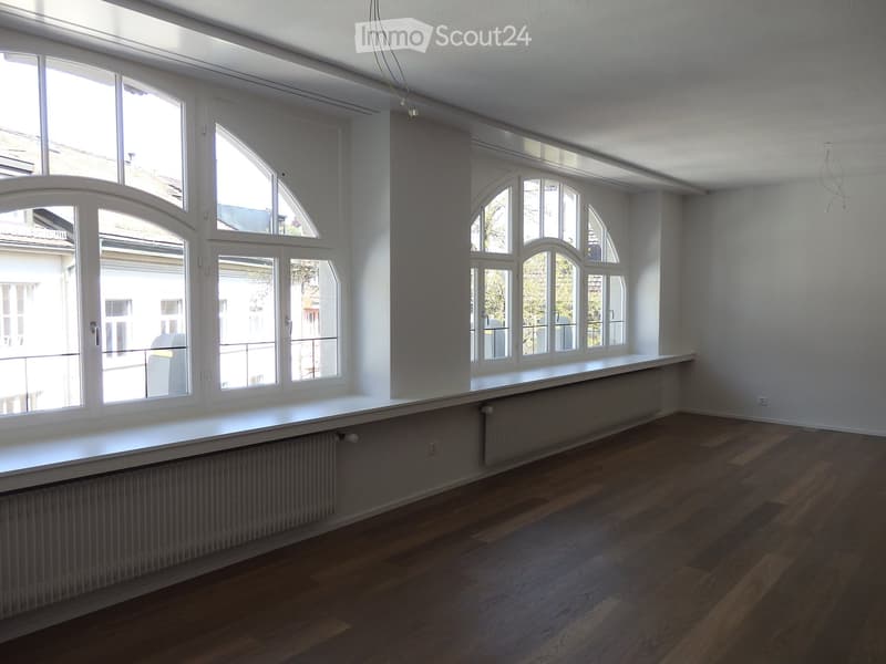 Empty room with wooden flooring, white walls, large windows, and radiators.