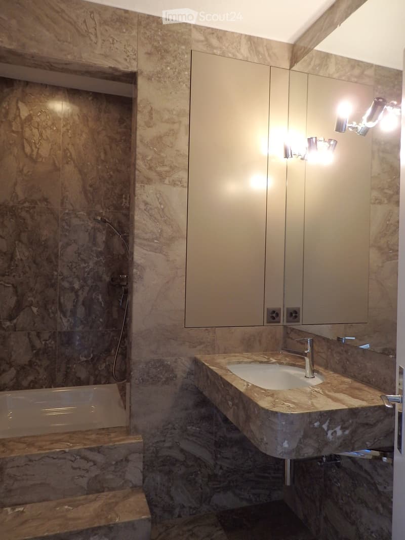 Modern bathroom with marble walls and floors, a large sink with a modern faucet, a mirrored cabinet, and a bathtub with a glass shower enclosure.