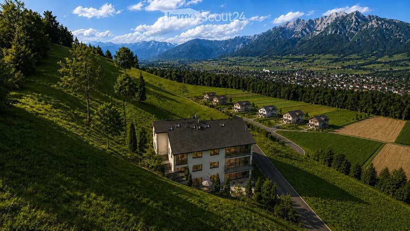 A modern two-story house with a dark roof and large windows, situated on a hillside with a panoramic view of a valley and mountains. The house features a balcony and is surrounded by green fields and trees.