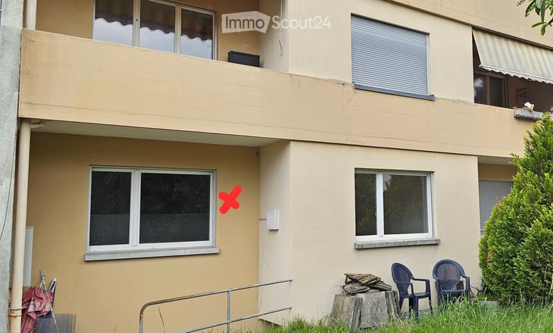Two-story residential building with beige exterior, multiple windows, and a small balcony area with outdoor seating.
