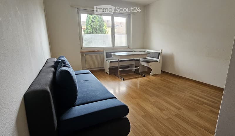 A small, well-lit room with wooden flooring, a blue sofa, a white table, and two benches. The room has large windows with a view of greenery outside.