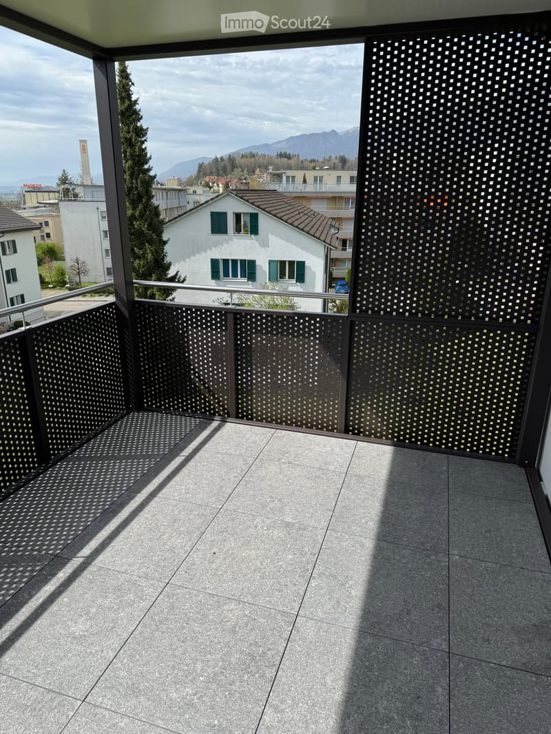 Balcony with grey tiled floor, metal railing, and decorative perforated metal panels, overlooking a cityscape with houses, trees, and distant mountains.
