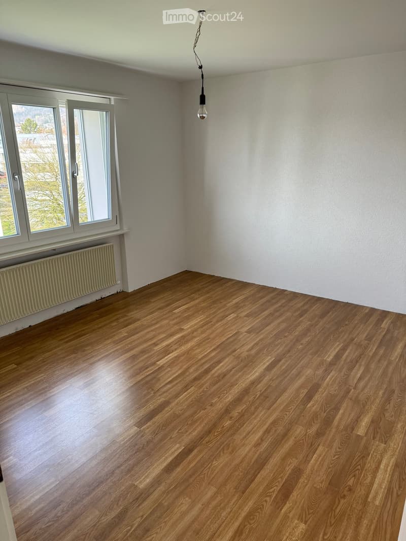 Empty room with wooden flooring, white walls, large windows, radiator, and a single hanging light fixture.