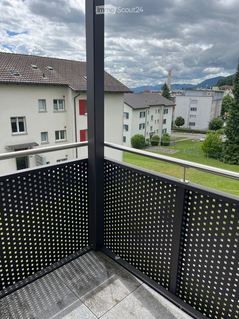 Balcony with black metal railing and patterned design, overlooking a residential area with houses, green lawn, and distant mountains.