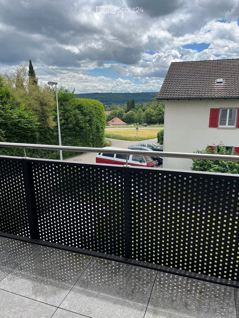 Balcony with a black lattice fence, metal railing, and tiled floor, overlooking a driveway with parked cars and a house with red shutters.
