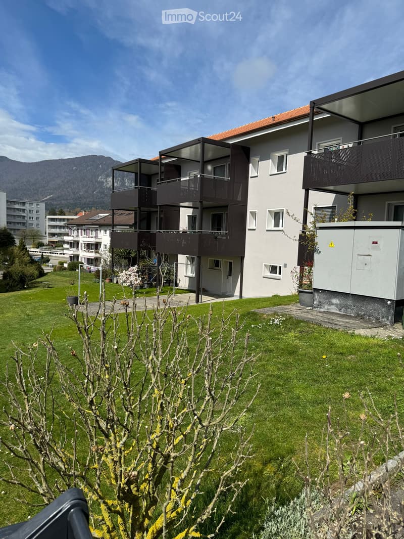 Modern multi-story residential building with balconies, surrounded by a well-maintained lawn and trees, with mountains in the background.
