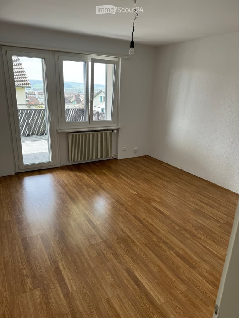 Empty room with wooden flooring, white walls, large windows, radiator, and a hanging light fixture.