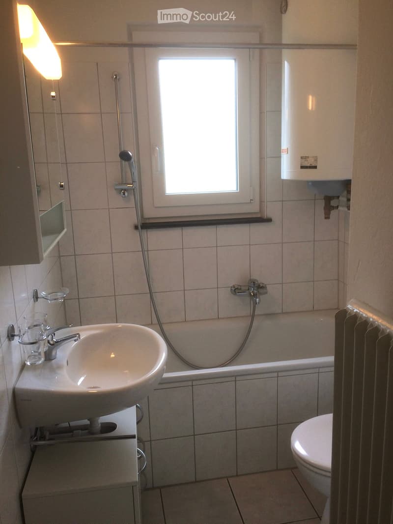 Modern bathroom with a sink, bathtub, toilet, and a window for natural light. Includes a mirror, cups on the sink, and a shower head.