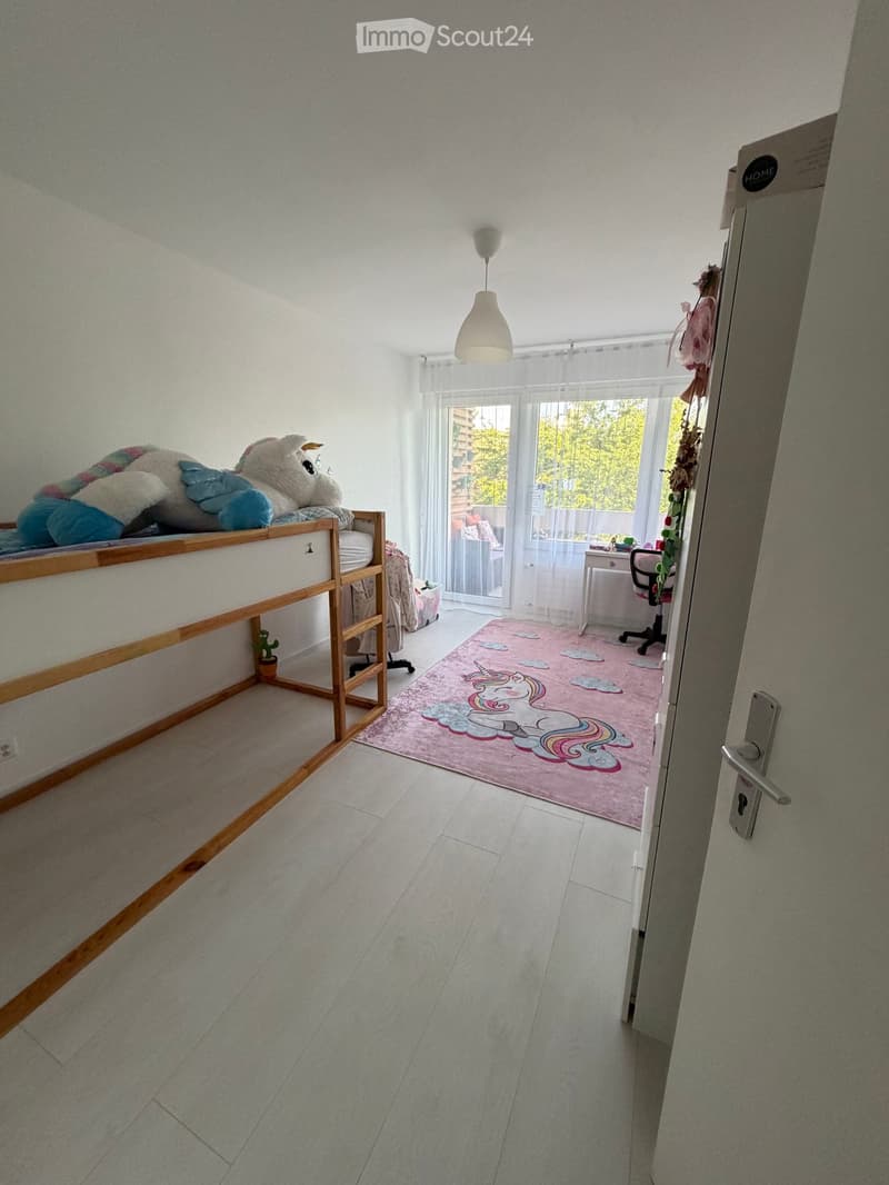A well-lit bedroom with a bunk bed, a pink rug with a unicorn design, a small table, and a chair near the window.