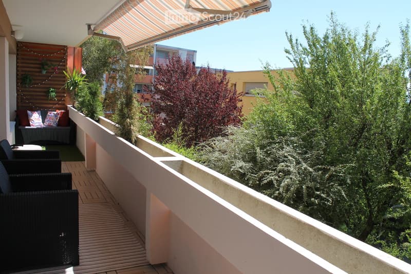 Balcony with wooden decking, black wicker chairs, small sofa with colorful cushions, and a view of greenery and neighboring buildings.