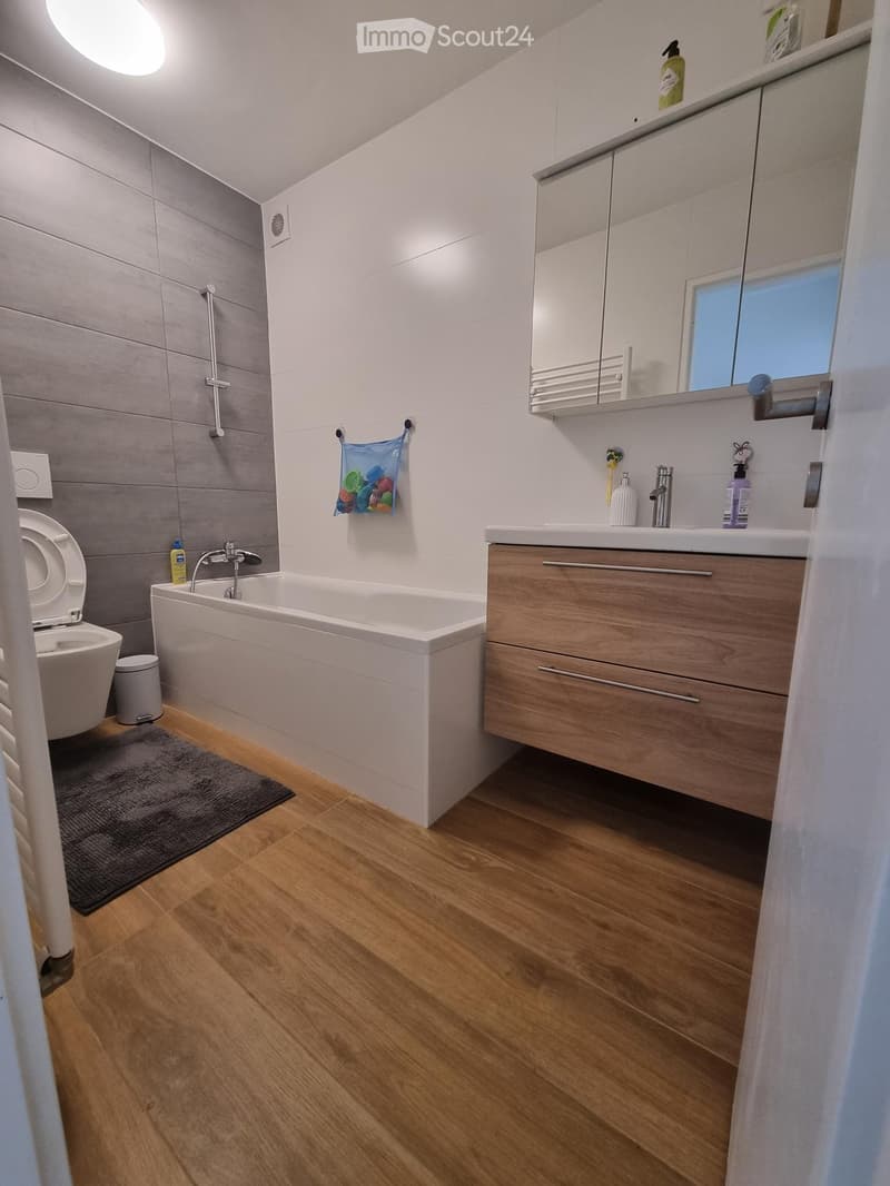 Modern bathroom with wooden flooring, a bathtub, toilet, sink with mirror, and a wooden cabinet. Includes a shower area with a handle and a small bag hanging on the wall.