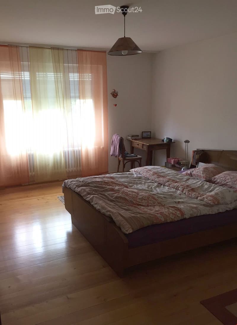 Bright bedroom with wooden flooring, a large bed with patterned bedding, a desk with a chair, and a hanging lamp.