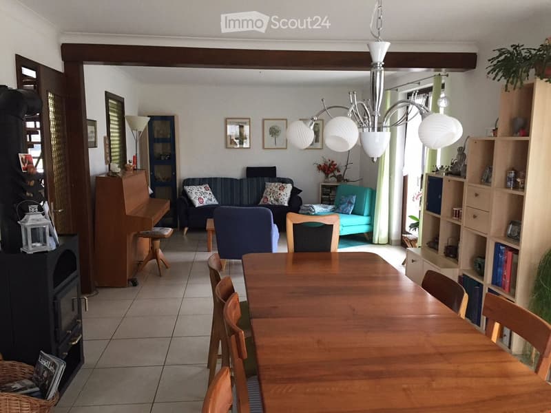 A well-lit living and dining area with a wooden dining table and chairs, a sofa, a piano, and a fireplace. The space features a chandelier, shelves, and decorative items.