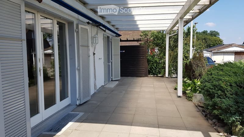 A covered patio area with a tiled floor, white wooden pergola, and glass doors leading to the house. The area is surrounded by greenery and has a small garden with plants and shrubs.