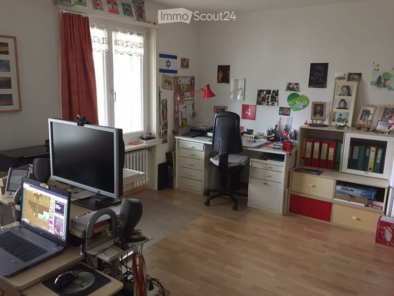 A well-lit home office with wooden flooring, a large window with red curtains, and various office equipment including a laptop, monitor, desk, chair, and filing cabinet.