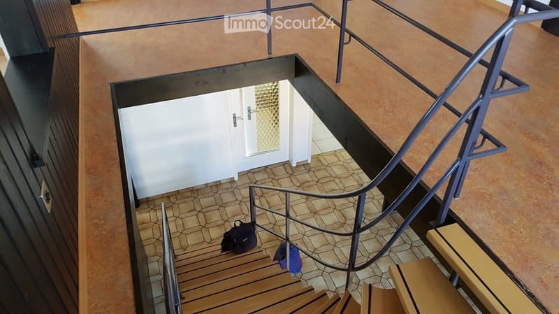 A modern hallway with a staircase leading to an upper level, featuring wooden steps, metal railings, and a hexagonal tile floor. A white door is visible at the end of the hallway.