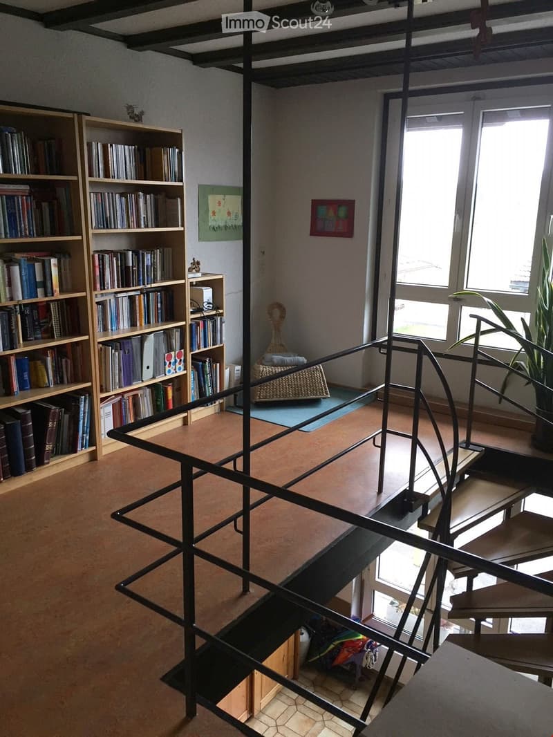 A modern hallway with wooden flooring, a staircase with metal railings, and a small seating area with a woven chair and a blue mat. Bookshelves line one wall, and there are windows allowing natural light to enter.