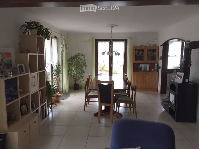 Bright living and dining area with tiled flooring, large windows, a dining table with chairs, a sofa, a bookshelf, and a fireplace.