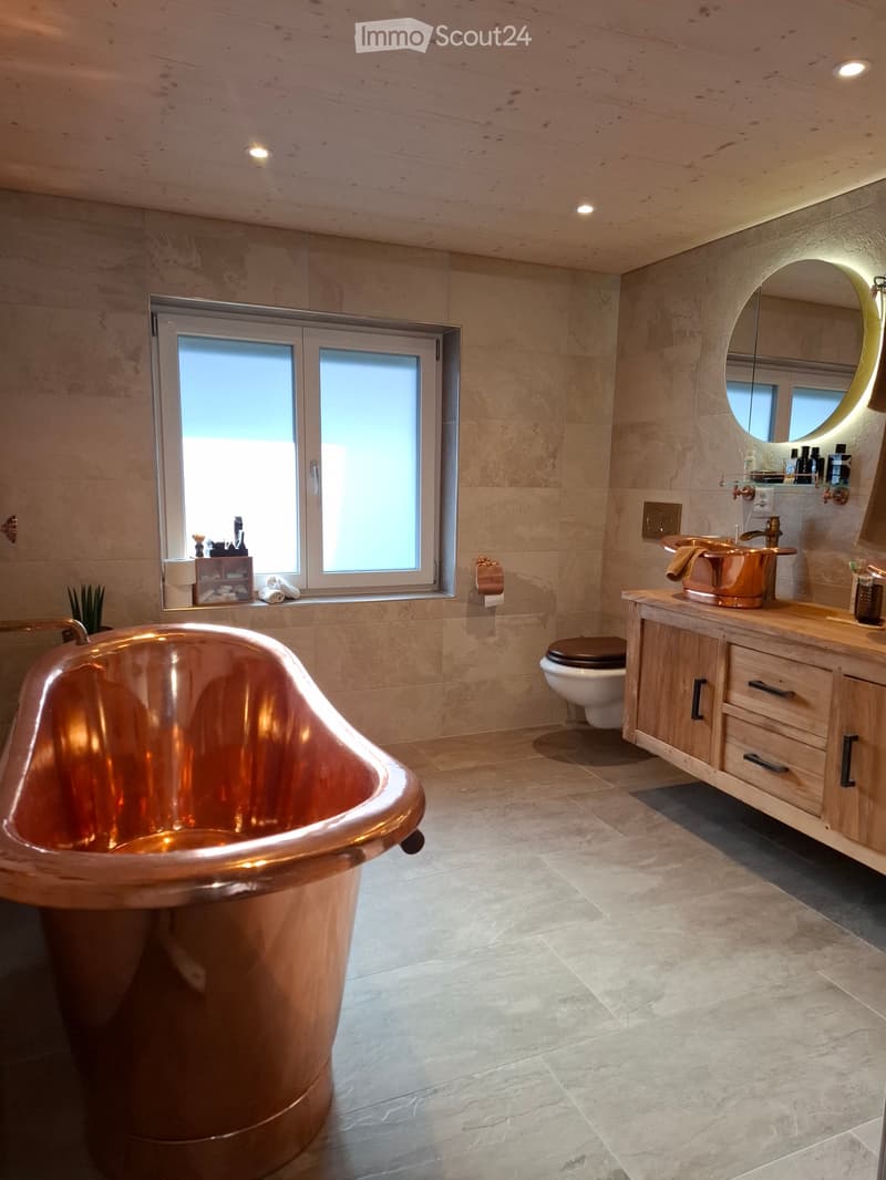 Modern bathroom with a large copper bathtub, marble tiles, a vanity with a sink, and a toilet. Features a large window with frosted glass and a round mirror with a gold frame.