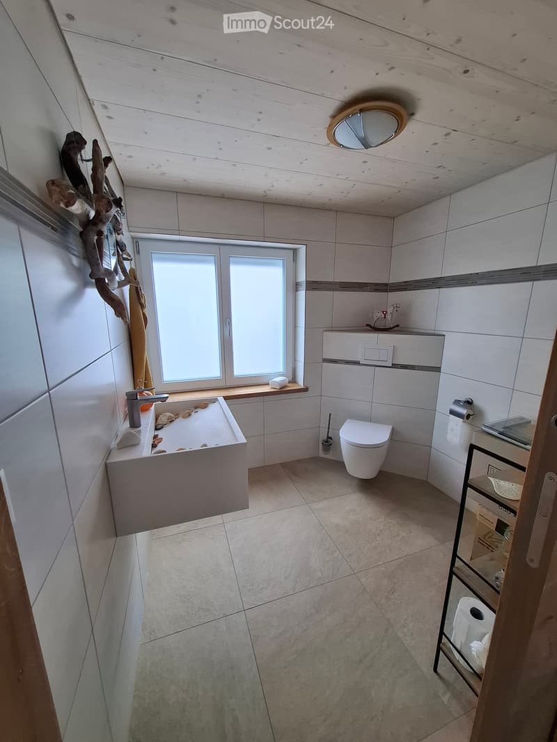 Modern bathroom with a large sink, toilet, and a window providing natural light. The room features white tiled walls, a wooden ceiling, and a small shelf with decorative items.