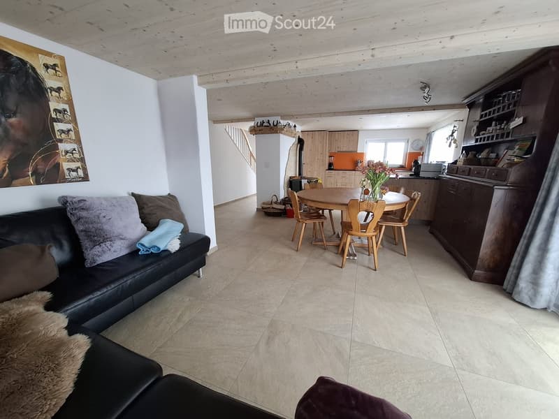 Open-plan living and dining area with a black leather sofa, wooden dining table with chairs, kitchen area with cabinets and appliances, and a staircase leading to another level.