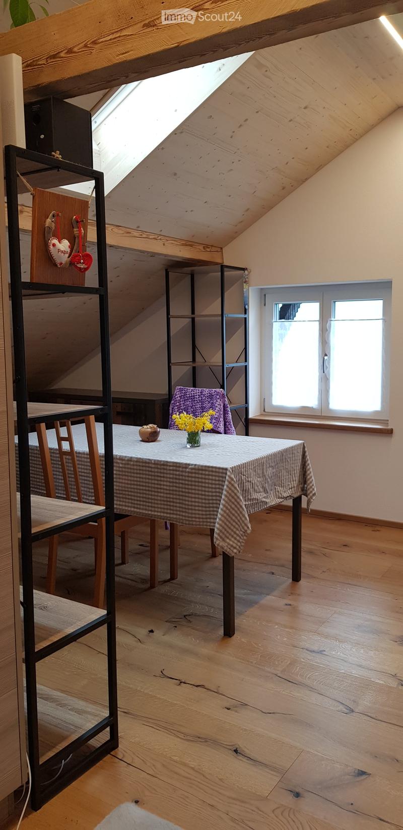 Bright attic room with wooden floors, slanted wooden ceiling, large windows, and a small table with a vase of yellow flowers.