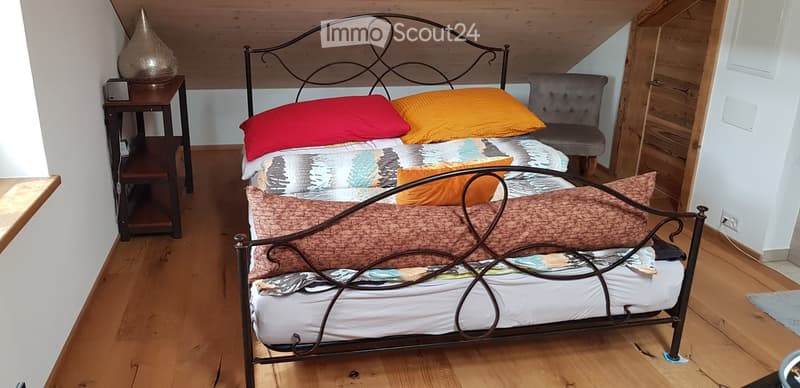 Cozy bedroom with wooden floors, a metal-framed bed with colorful bedding, a nightstand with a decorative vase, and a small armchair.