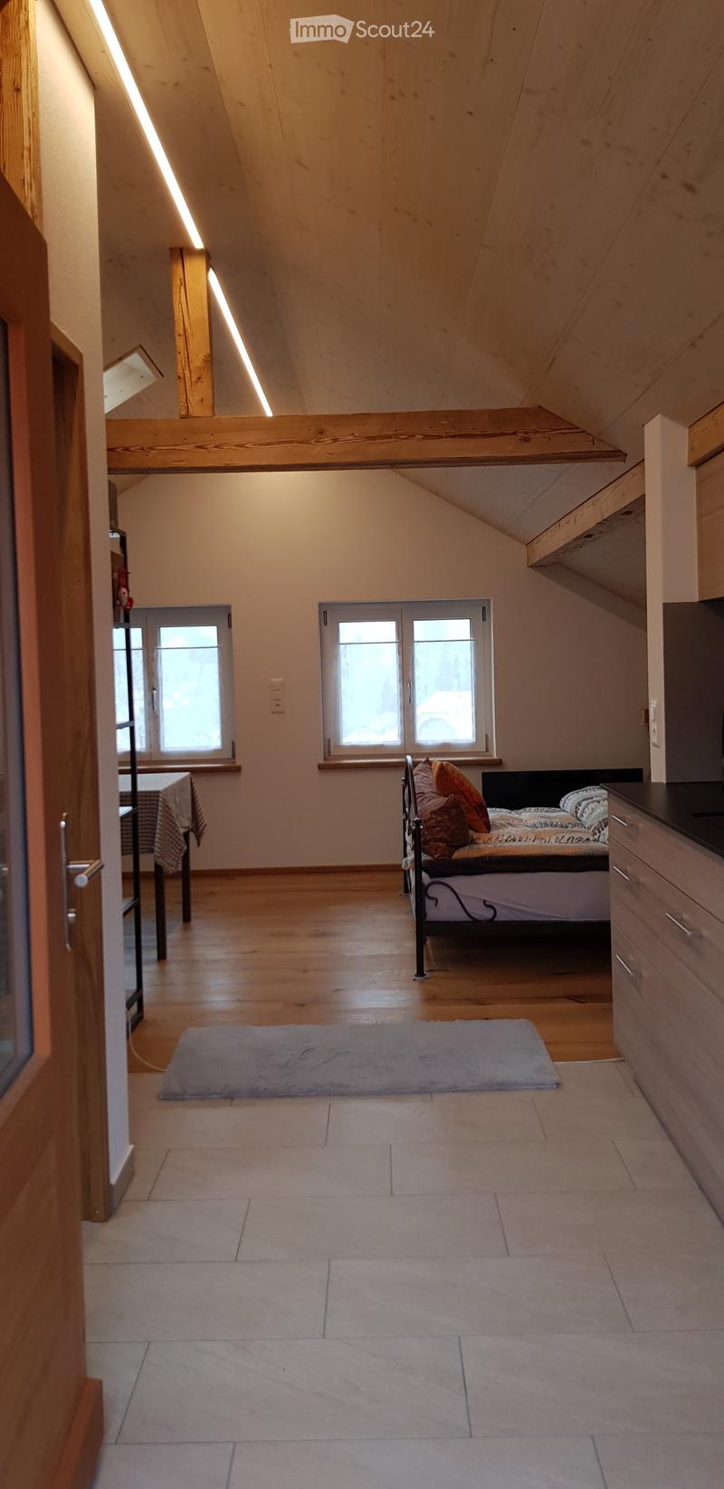 Bright attic bedroom with wooden beams, exposed wooden ceiling, large windows, bed with pillows, nightstand, small table with chair, and a gray rug.