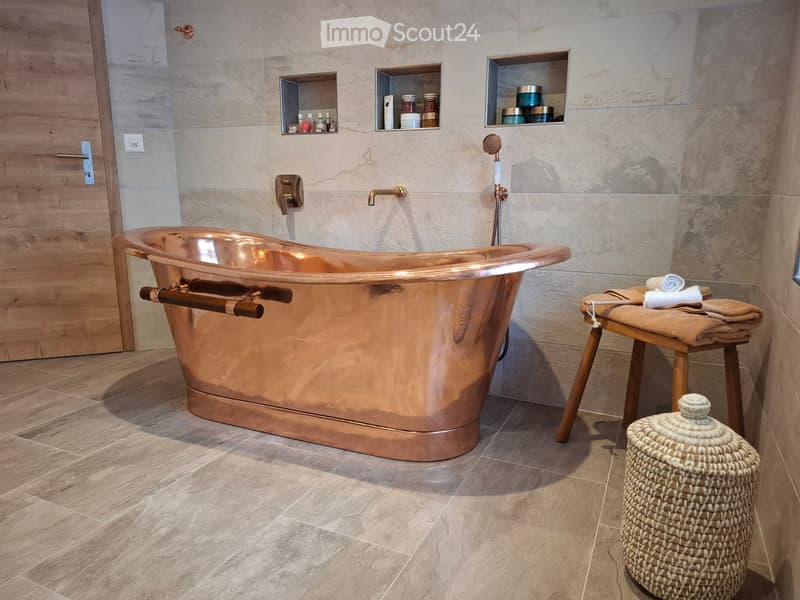 Modern bathroom with a copper bathtub, marble walls, wooden floor, towel on a stool, and a wicker basket.