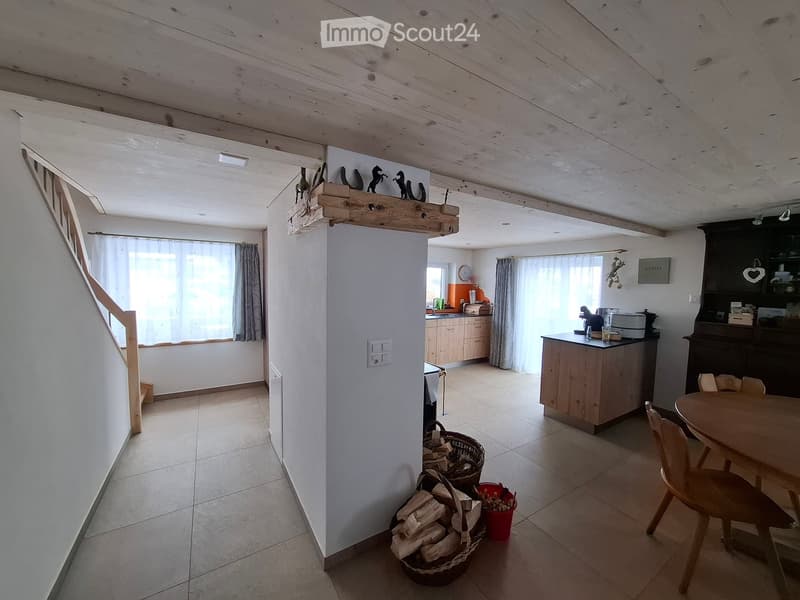 Open-plan living and dining area with wooden beams, tiled floor, and a staircase leading to an upper level. Includes a dining table with chairs, a kitchen area with cabinets and a counter, and baskets with firewood.