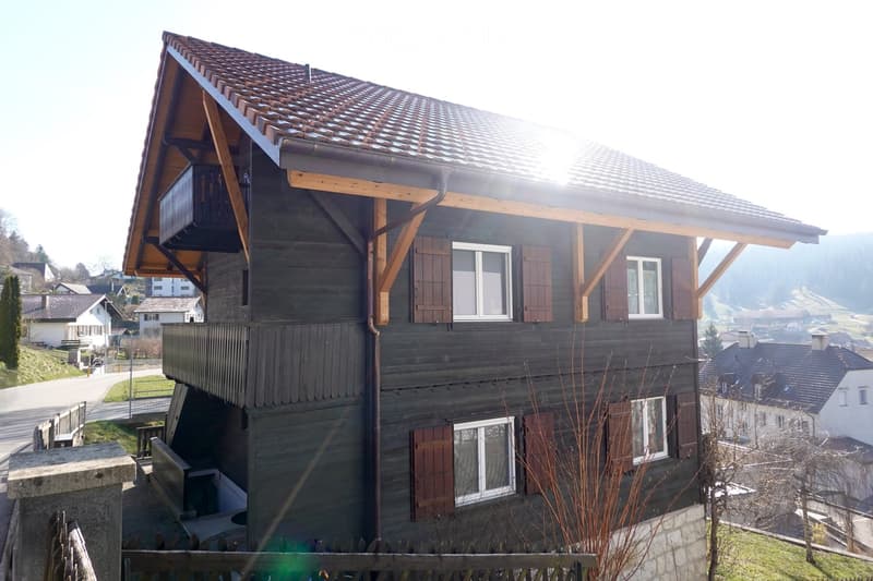 A two-story wooden house with a tiled roof, multiple windows with shutters, and a small balcony on the upper level. The house is situated in a rural area with other houses and greenery in the background.