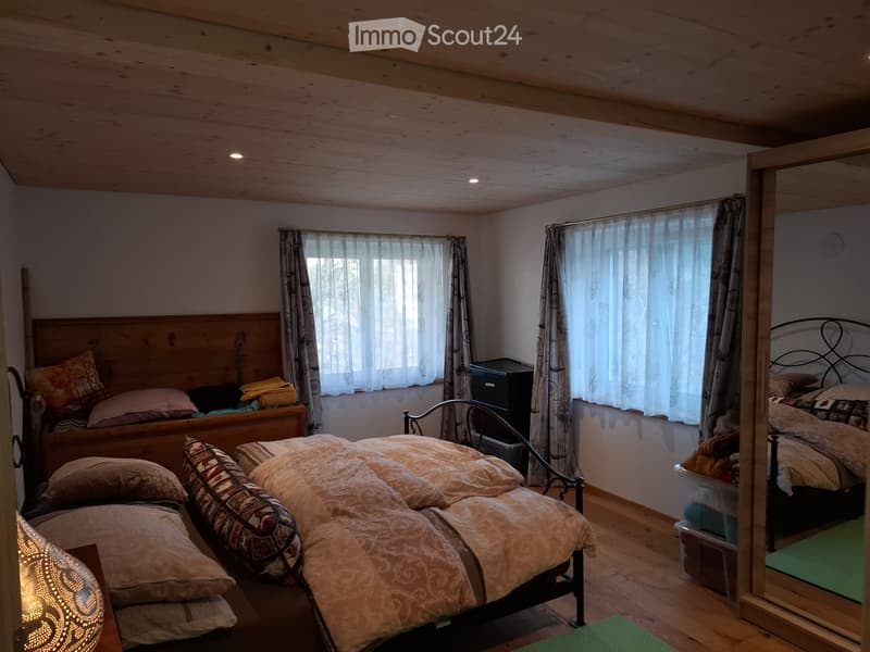 Cozy bedroom with wooden ceiling, two beds, nightstands, and a mirror. Light-colored bedding and pillows, with curtains on windows.