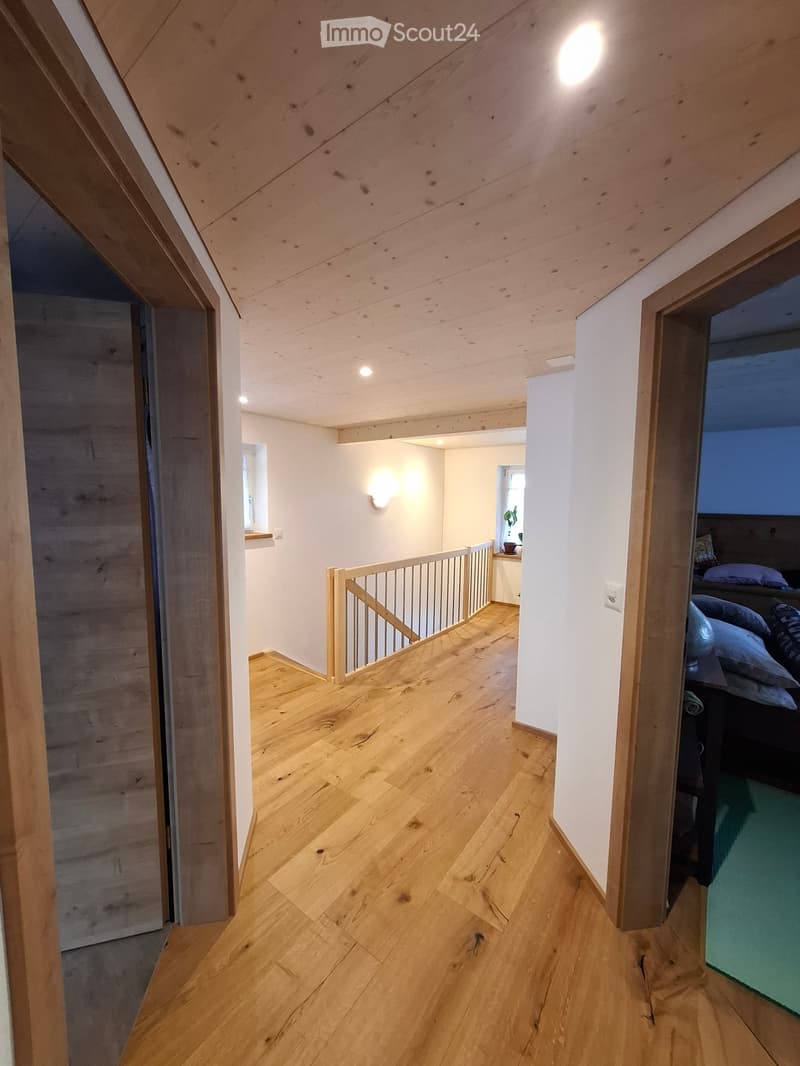 Bright, modern hallway with wooden flooring, white walls, and exposed wooden beams on the ceiling. Features a staircase with a wooden railing leading to an upper level, and a glimpse of a bedroom with a bed and pillows visible at the end of the hallway.