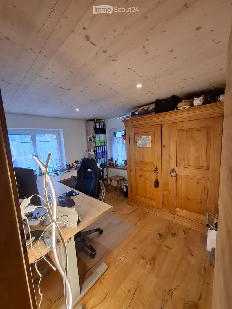 A well-lit, wooden-floored room with a modern desk setup, a comfortable chair, and a large wooden cabinet. The room features large windows with curtains, a small stool, and a bookshelf with binders and books.