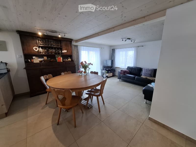 Open-plan living and dining area with a round wooden dining table, six chairs, a black leather sofa, a wooden cabinet, and a television on a stand.