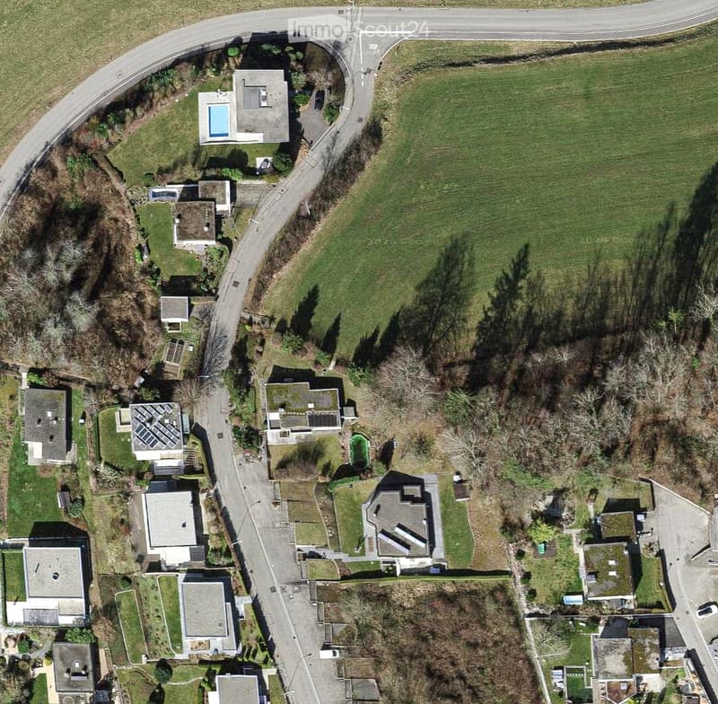 Aerial view of a residential area with multiple houses, green lawns, and a swimming pool visible in the upper left area.