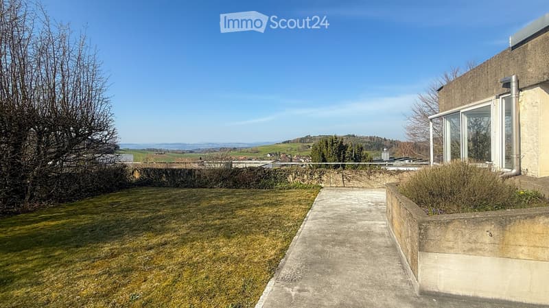 A raised garden area with a concrete pathway leading to a modern building with large windows, surrounded by a grassy lawn and distant countryside views.