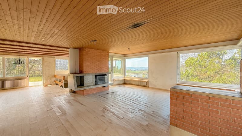 Bright, open living space with wooden floors, exposed wooden ceiling beams, large windows offering scenic views, and a central brick fireplace.