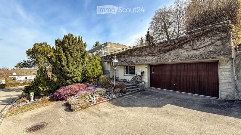 Modern two-story house with a garage, surrounded by greenery and a landscaped garden. The house features a raised ground floor and a balcony on the upper level.