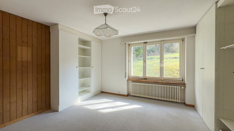 Empty room with white walls, carpeted floor, built-in white shelving unit, large window with radiator below, and a hanging light fixture.