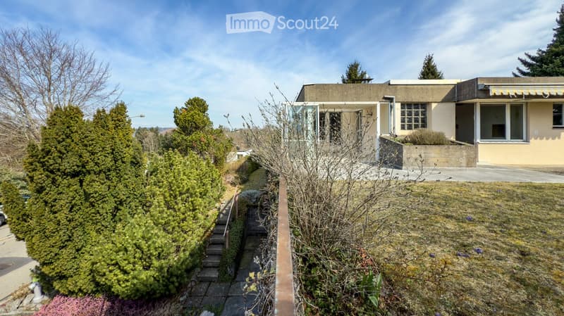 Two-story house with a raised ground floor, surrounded by greenery and a concrete pathway leading to the entrance.