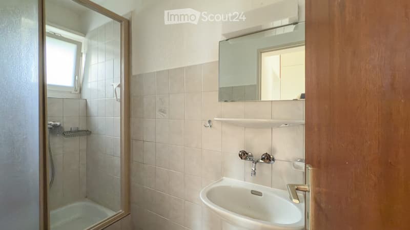 Bathroom with a sink, mirror, and a glass-enclosed shower. The walls are tiled in light beige, and the floor is also tiled.