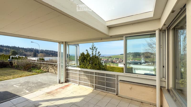 Bright, spacious balcony with large glass windows and sliding doors, offering scenic views of the countryside and distant hills.