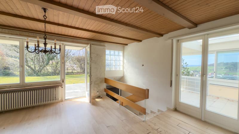 Bright, empty room with wooden flooring, white walls, large windows, and a wooden staircase leading to a balcony. The room features a chandelier and a radiator, with a view of a green landscape through the windows.