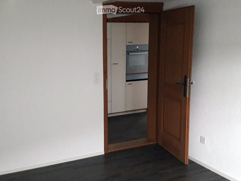 Unfurnished Room. Empty room with a wooden door leading to a kitchen area. The kitchen features white cabinets and a built-in oven.